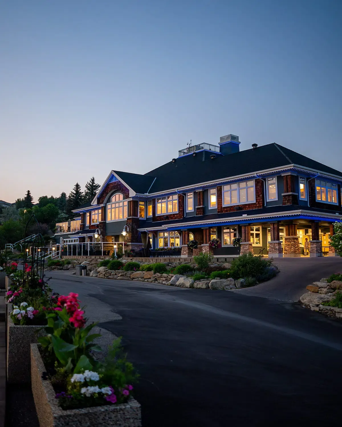 Lodge-style commercial building at dusk with lights along its rooflines and flower planters by the drive