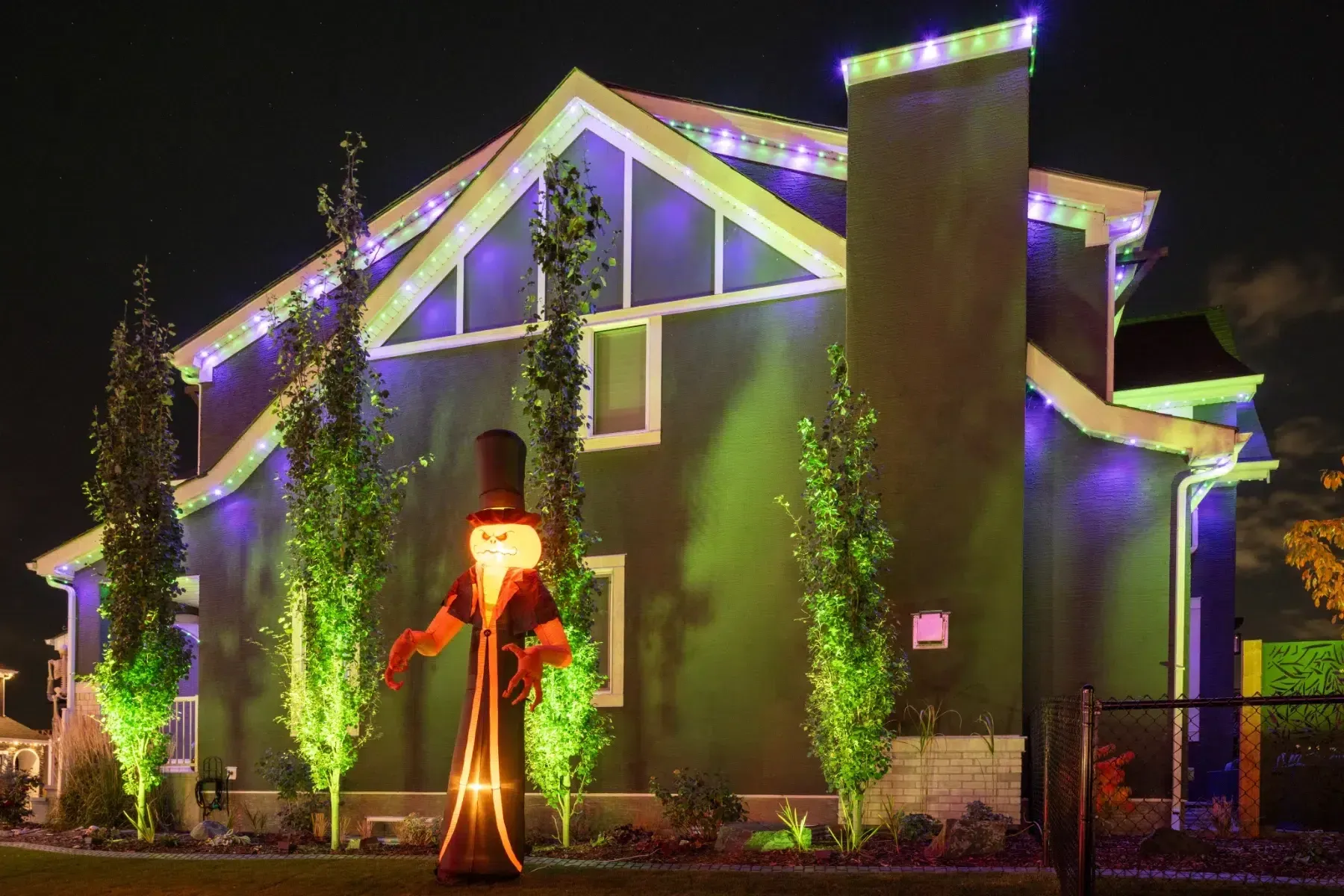 House with white roofline and green uplit trees beside a tall pumpkin figure