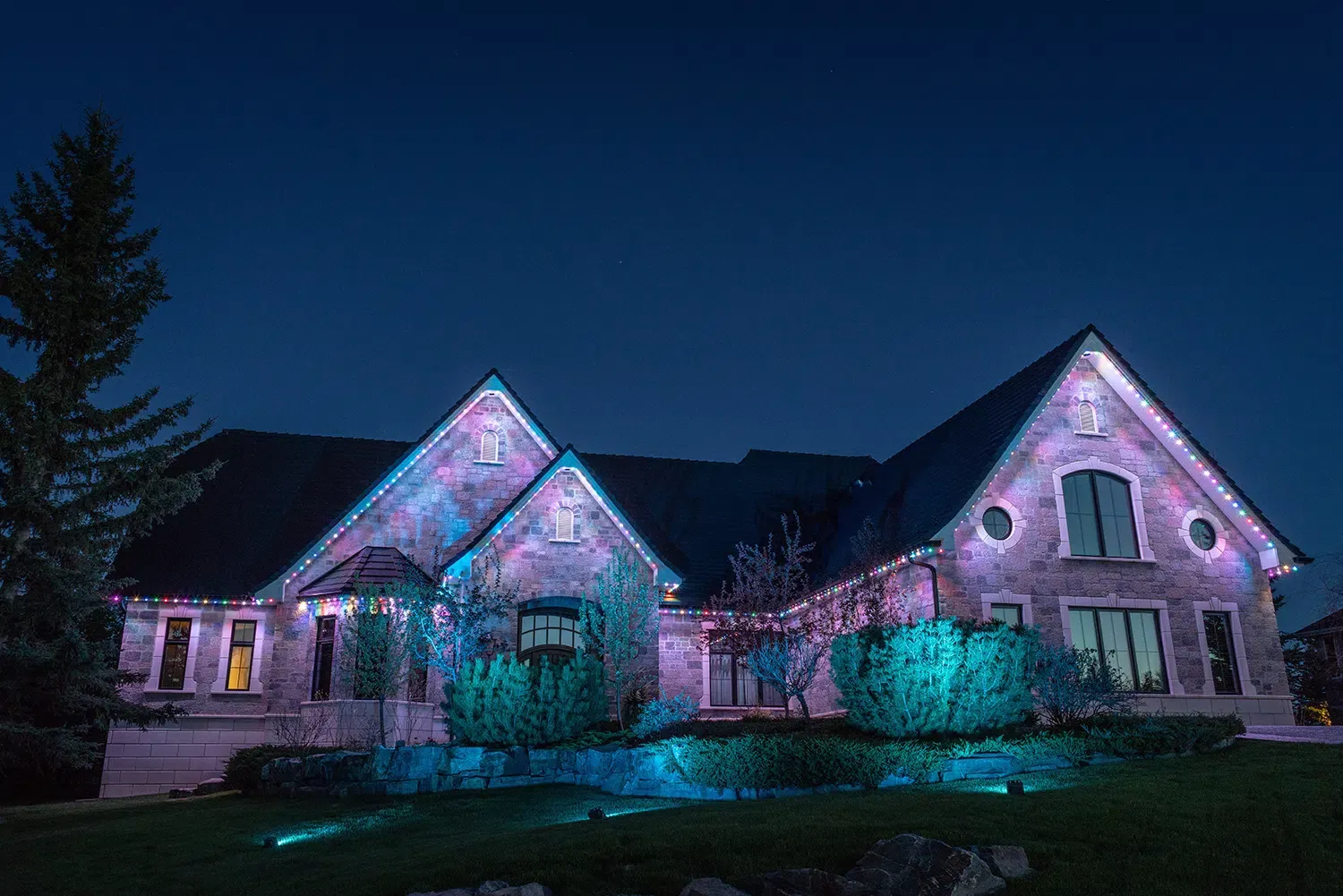 Stone house at dusk with teal uplights on the shrubs