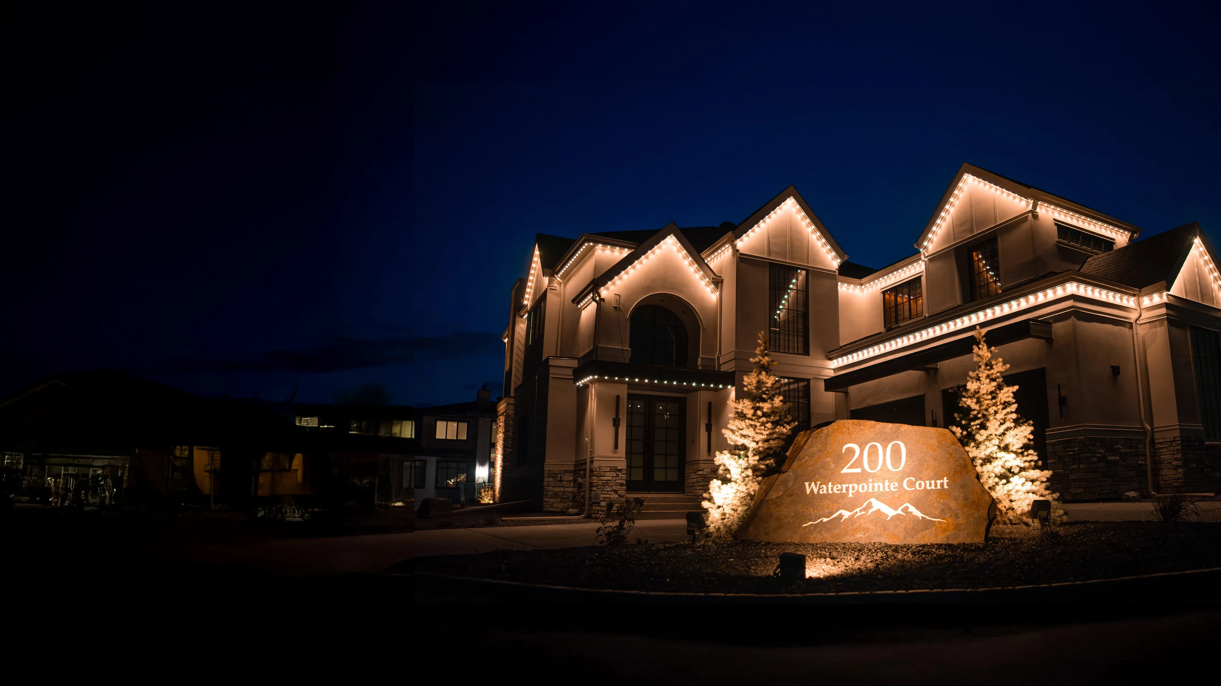 Large house at night with warm roofline lights and uplit evergreen trees beside a stone entrance sign