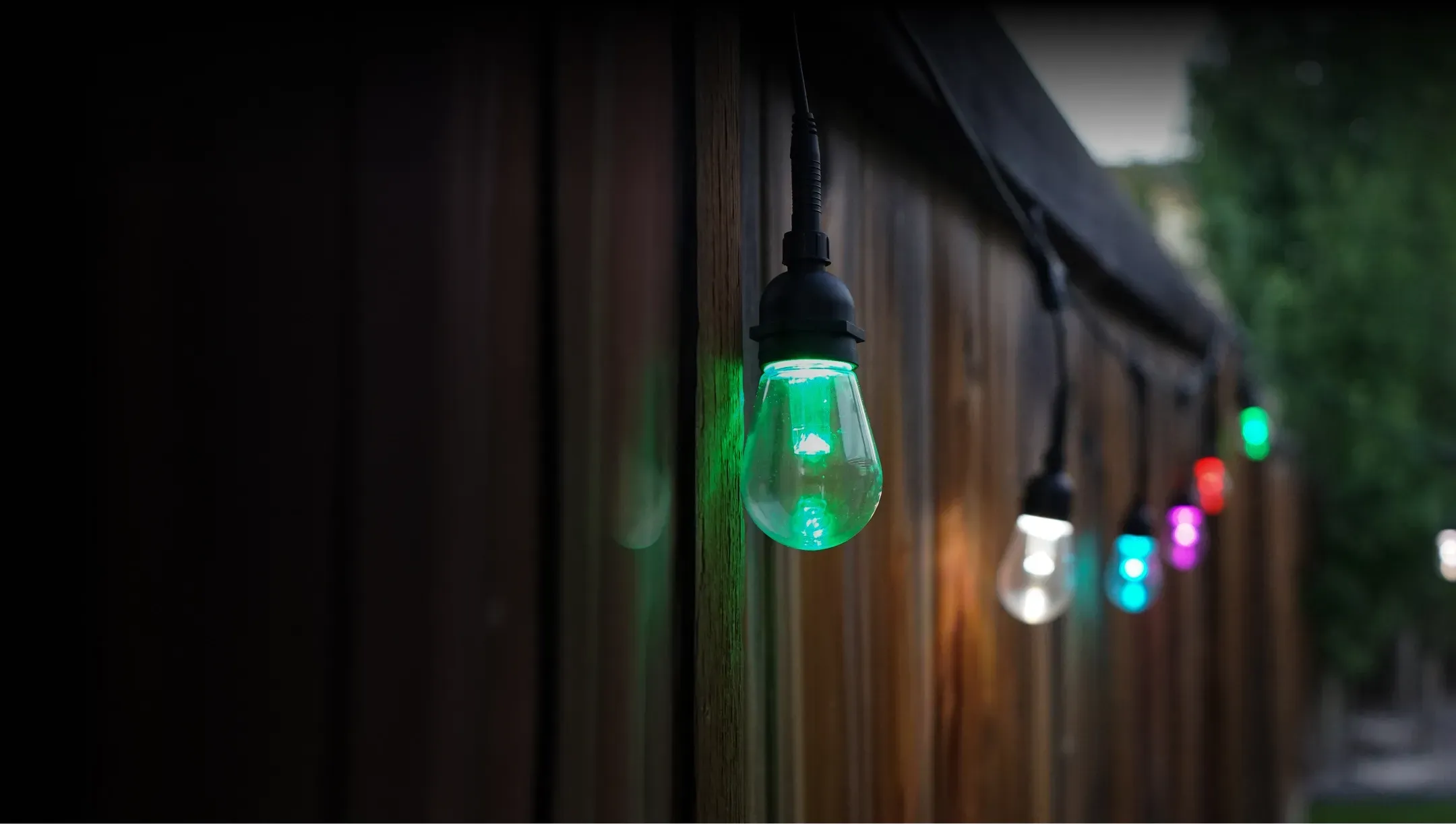 Row of smart string light bulbs glowing in different colours along a fence at dusk