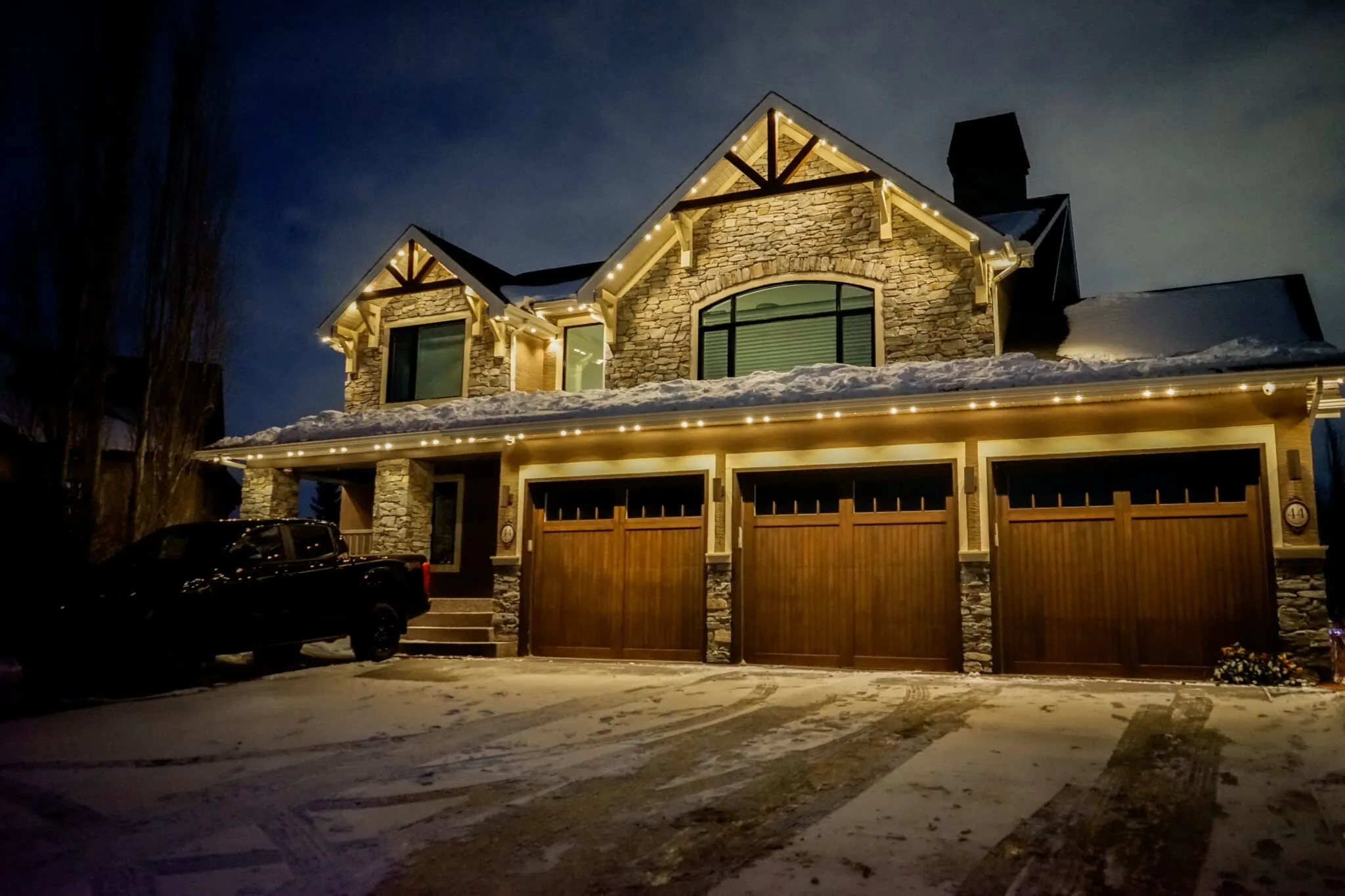 Stone house with a triple garage and warm white roofline lights on a snowy night